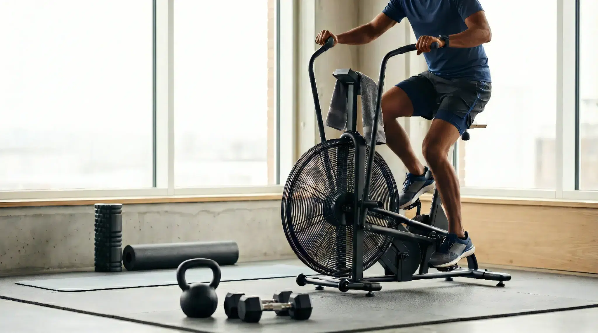 Cross-training for running using an air bike in a gym with kettlebell, dumbbells, foam roller and yoga mat for strength and recovery.